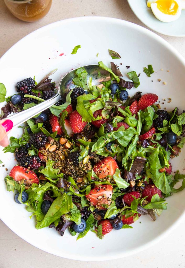 a berry salad being tossed with nuts and crispy quinoa in a large white serving bowl