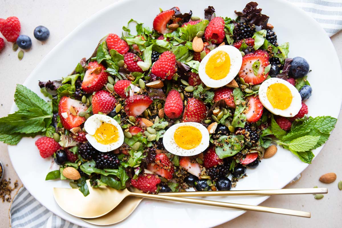 a large white platter filled with berries and salad greens and topped with soft-boiled eggs