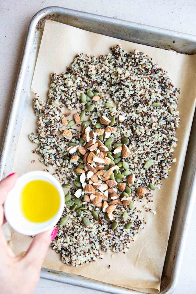 olive oil being poured on a baking tray filled with cooked quinoa, nuts and seeds