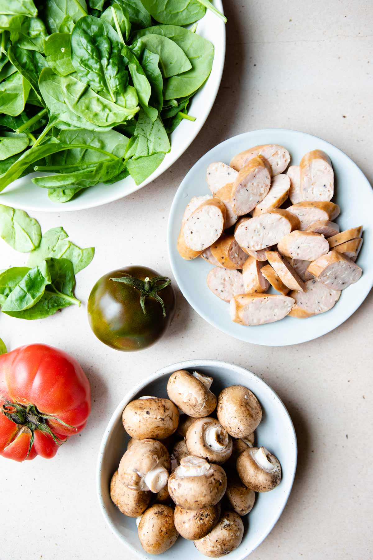 uncooked sausages sliced, raw onions, tomatoes and spinach on a beige surface