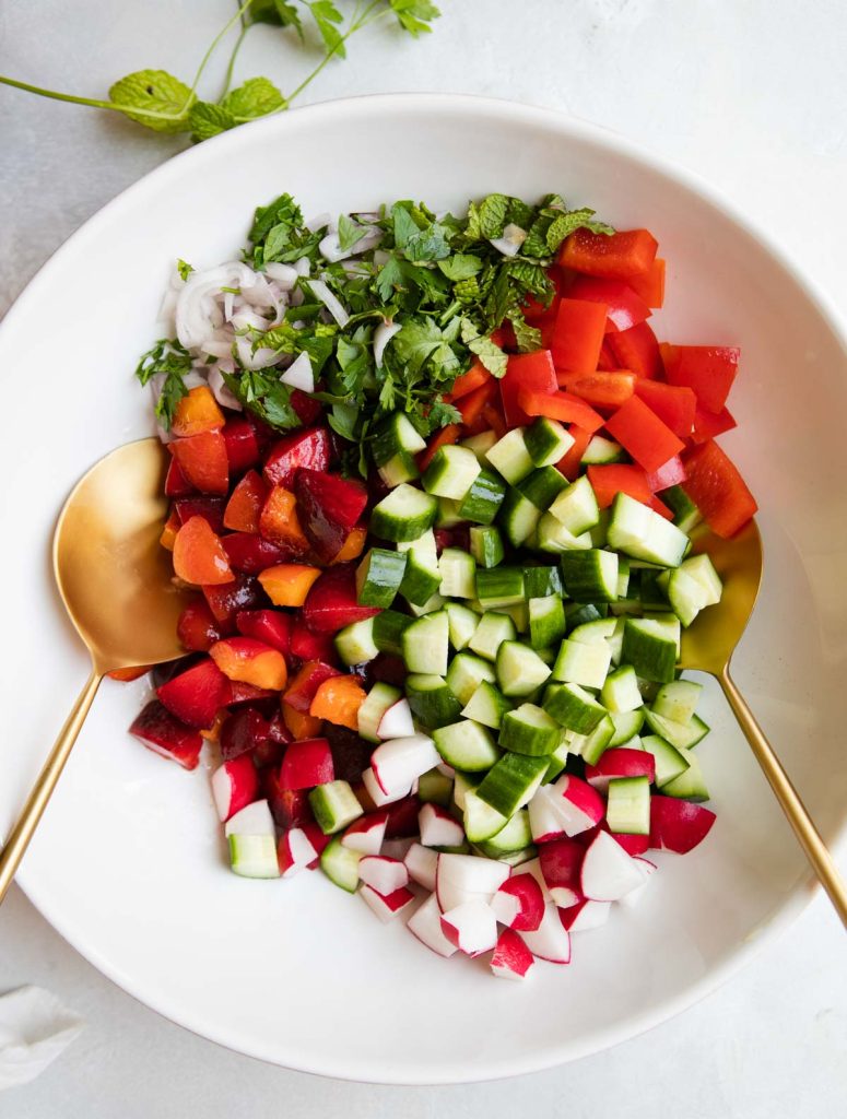 chopped up red peppers, cucumbers, radishes, herbs and plums and apricots in a white mixing bowl