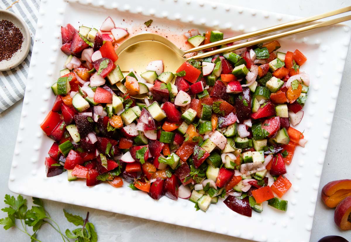 white platter filled with a veggie and stone fruit salad served with gold tongs