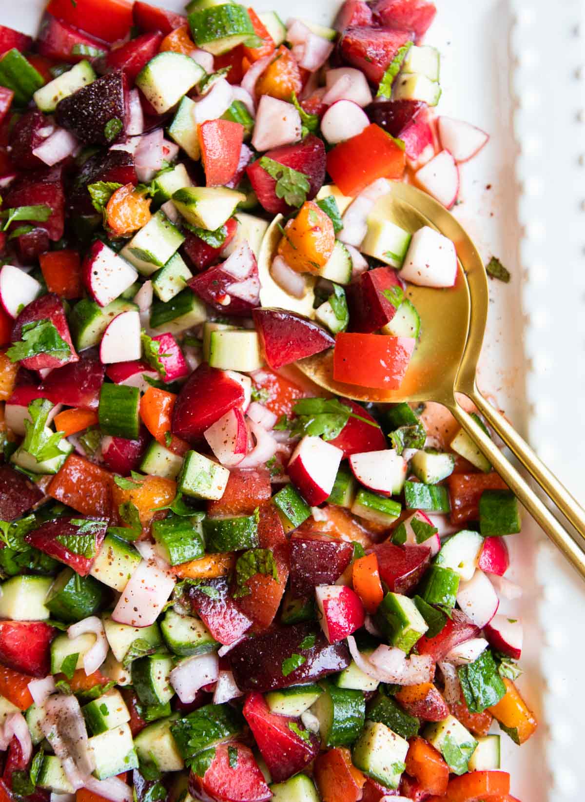 gold tongs scooping up a stone fruit salad with veggies and herbs mixed in