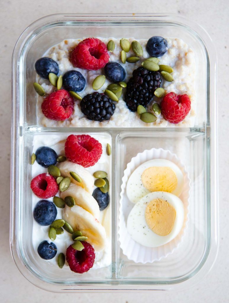 oatmeal and greek yogurt topped with berries and bananas and hardboiled eggs in a glass meal prep container 