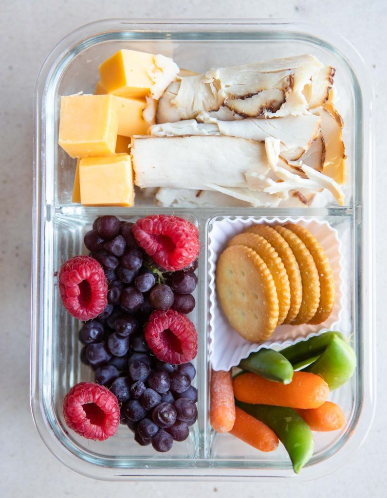 deli turkey, cubed cheddar cheese, grapes, raspberries and carrots and crackers in a glass meal prep container