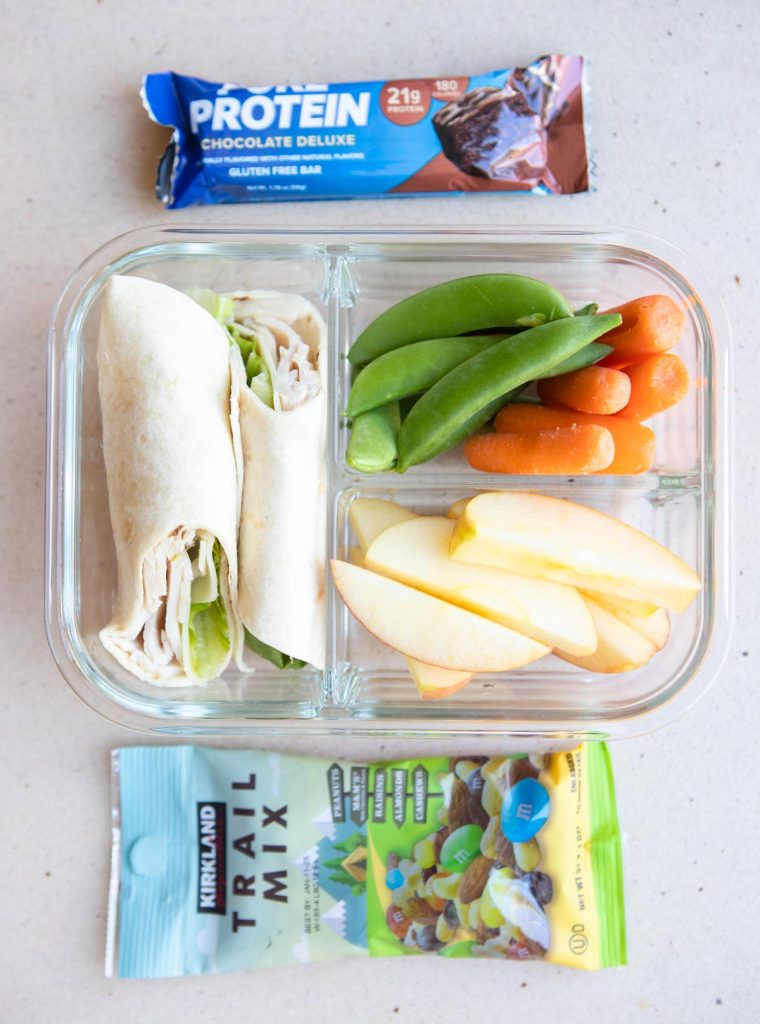 glass meal prep container with a turkey sandwich wrap, apples, carrots and snap peas along with a protein bar and trail mix