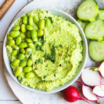 beige bowl filled with nutritious and delicious bright green edamame hummus with cucumbers and radishes served on the side