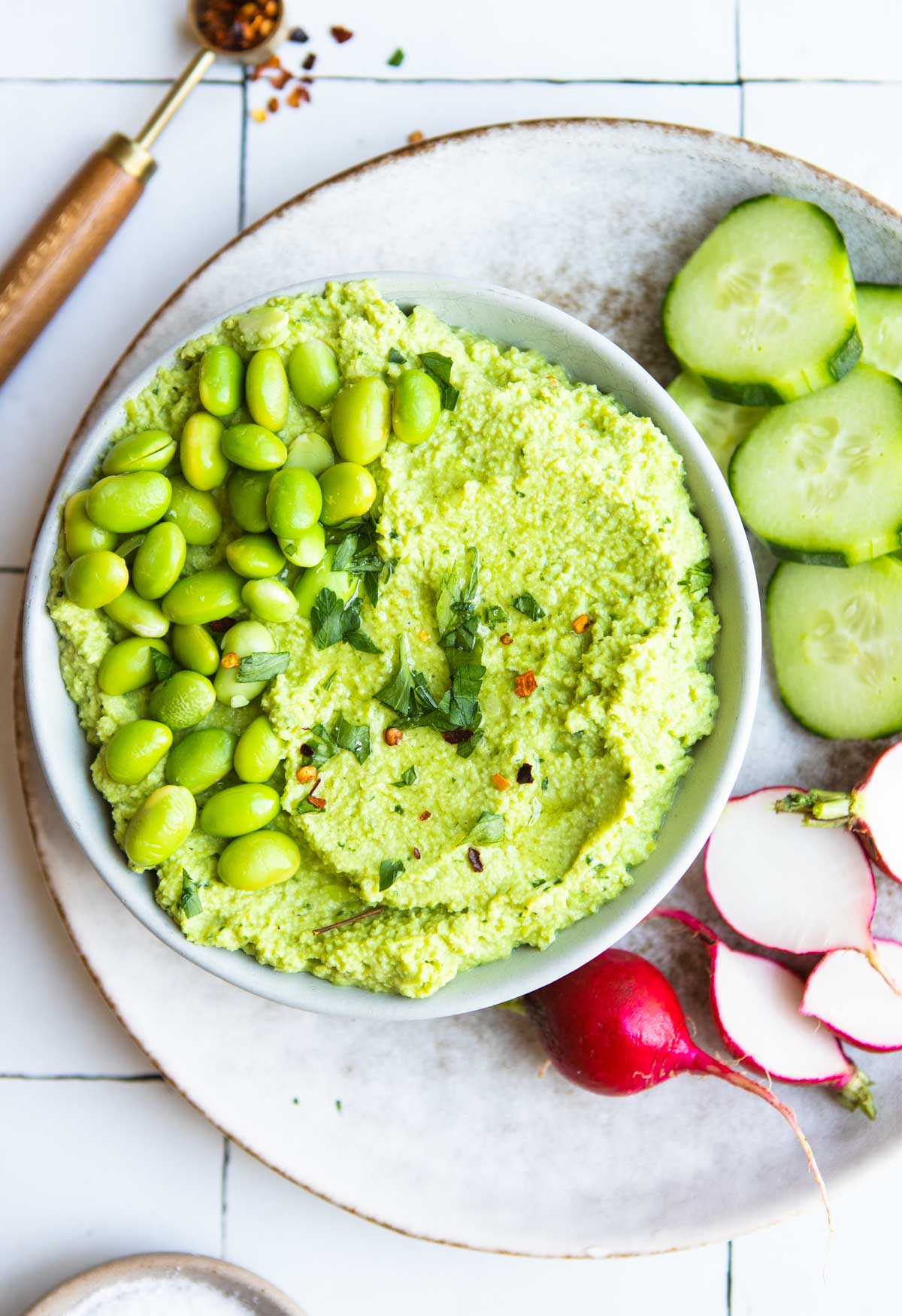 a gray bowl filled with edamame hummus and slices of cucumber and radish on the side