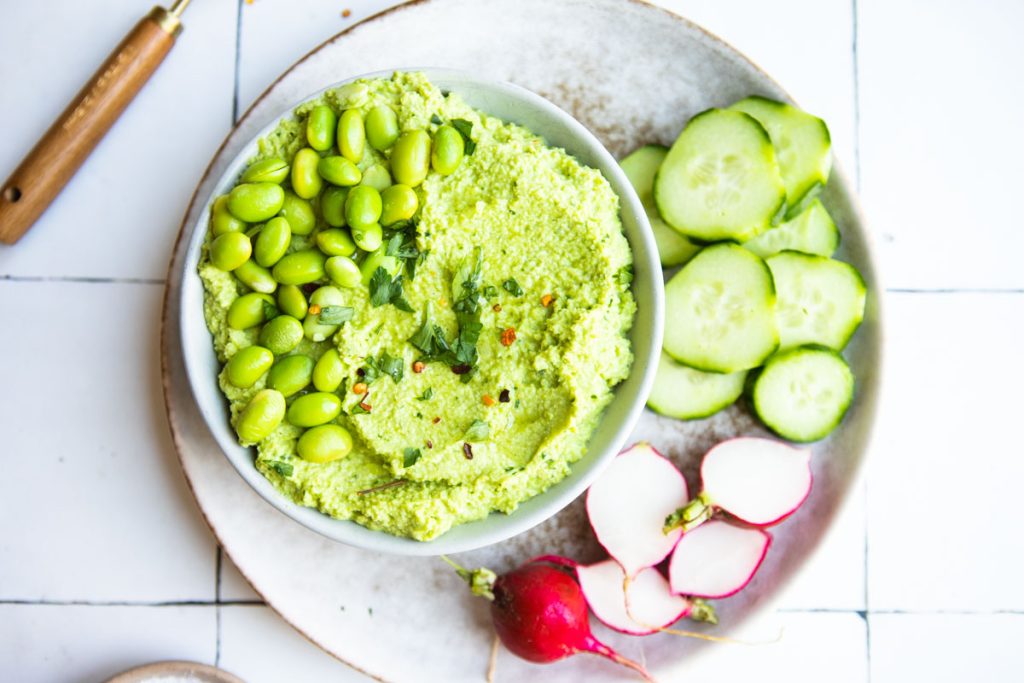 fresh edamame hummus in a gray bowl sitting next to a plate of sliced cucumbers and radishes