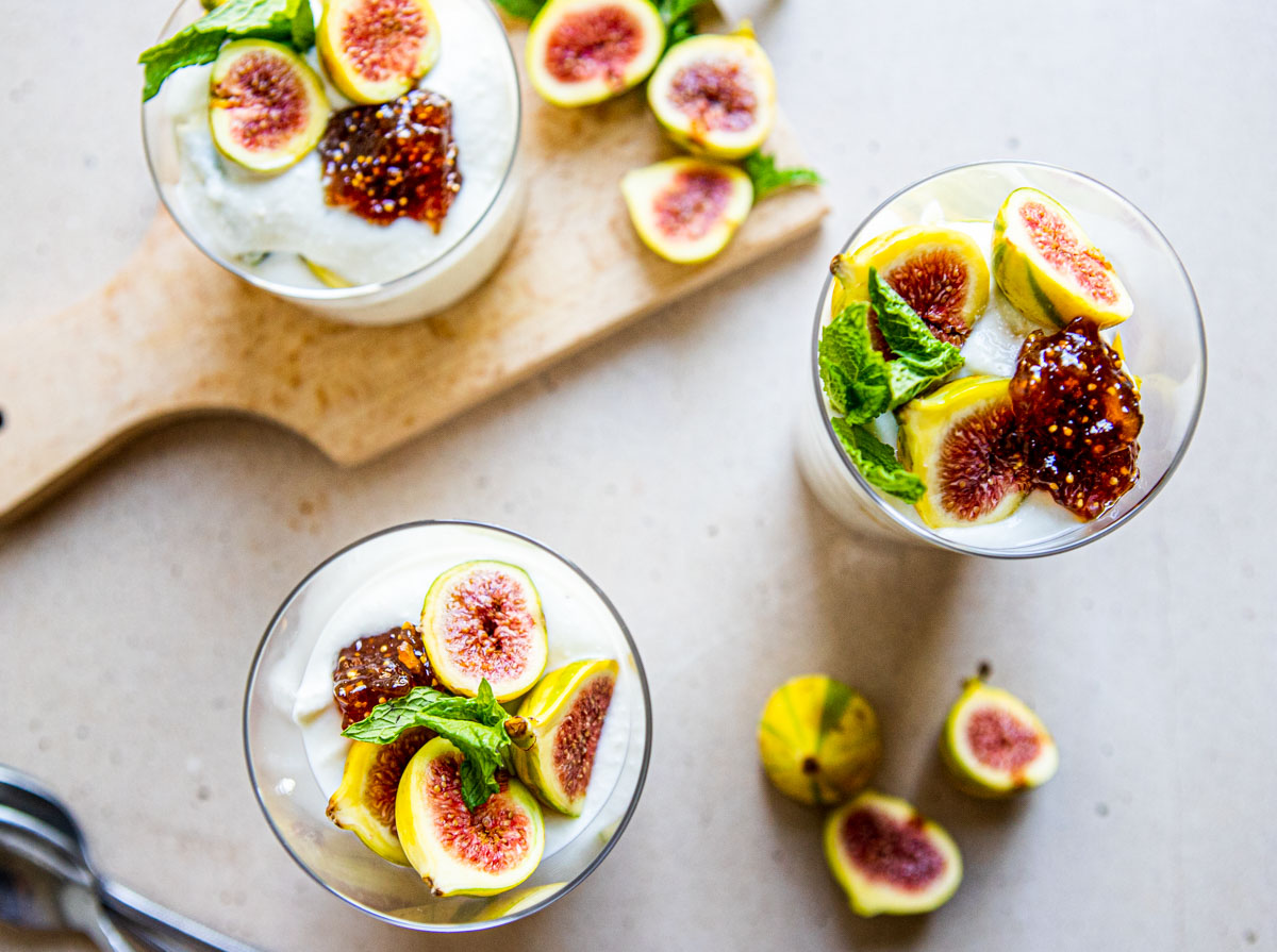 a table covered in fresh figs and 3 glasses filled with a ricotta cheese dessert and topped with fresh figs