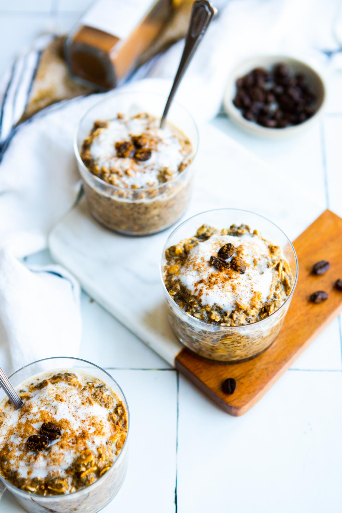 overnight coffee oats in 3 small glass cups topped with cold frothed milk and espresso beans