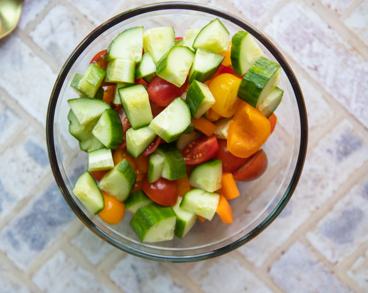 fresh diced cucumbers, peppers and halved cherry tomatoes in a glass bowl with olive oil and vinegar tossed in