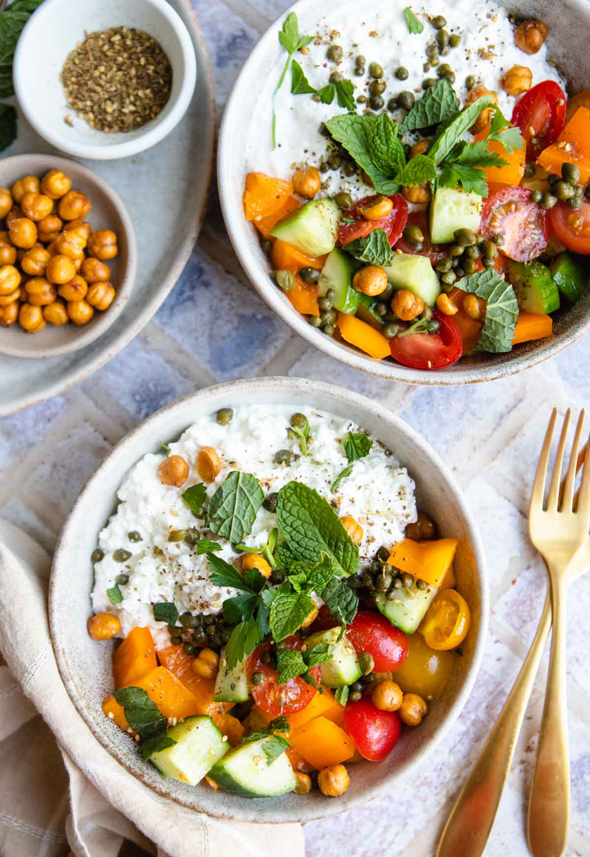 2 beige bowls filled with cottage cheese and a savory tomato and cucumber herbed salad, and topped with crispy chickpeas and seasoning