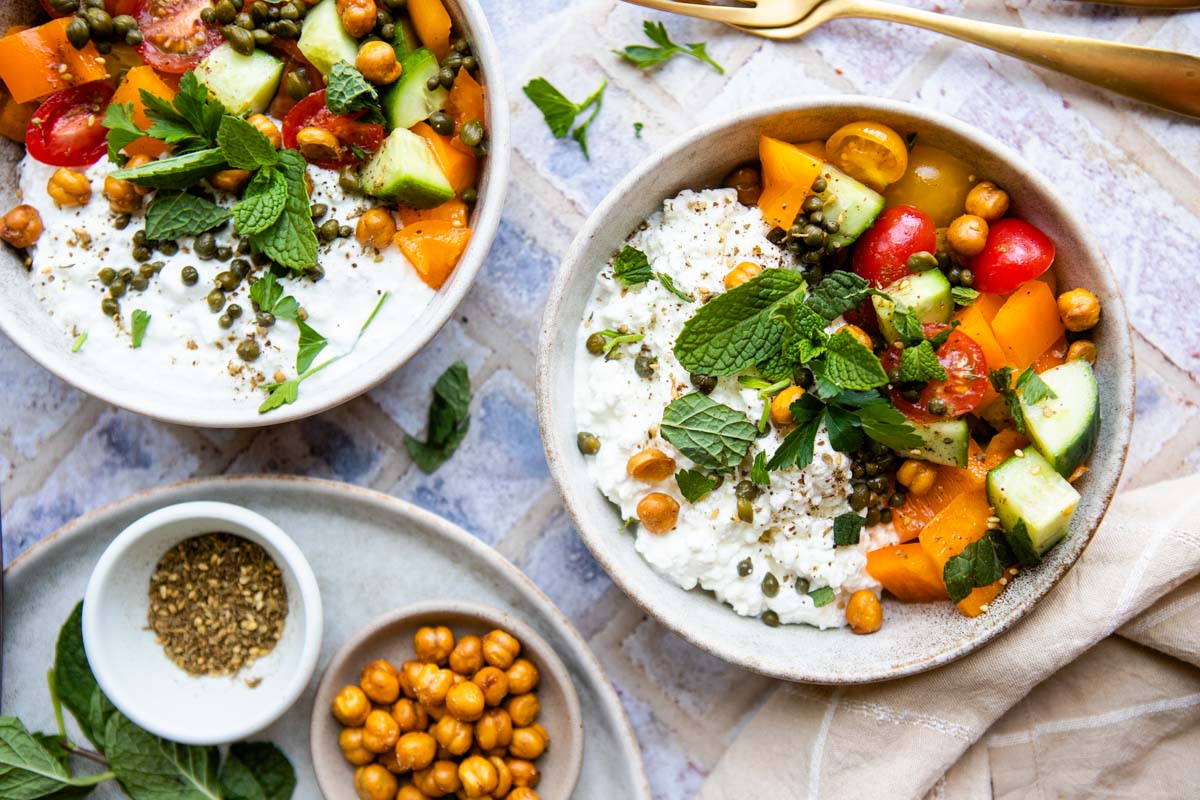 2 dished out savory cottage cheese bowls served in rustic ceramic bowls and the cottage cheese is garnished with mint and parsley and crispy chickpeas