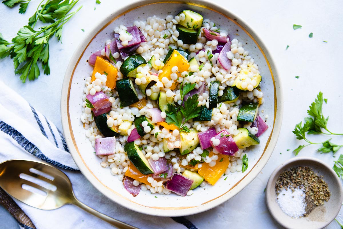 gold utensils next to a beige and gold rimmed plate filled with pearl couscous with vegetables