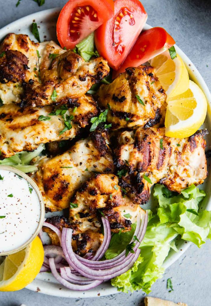 grilled greek yogurt chicken thighs on a plate with sliced tomatoes, red onions, lemons and a garlic greek yogurt sauce